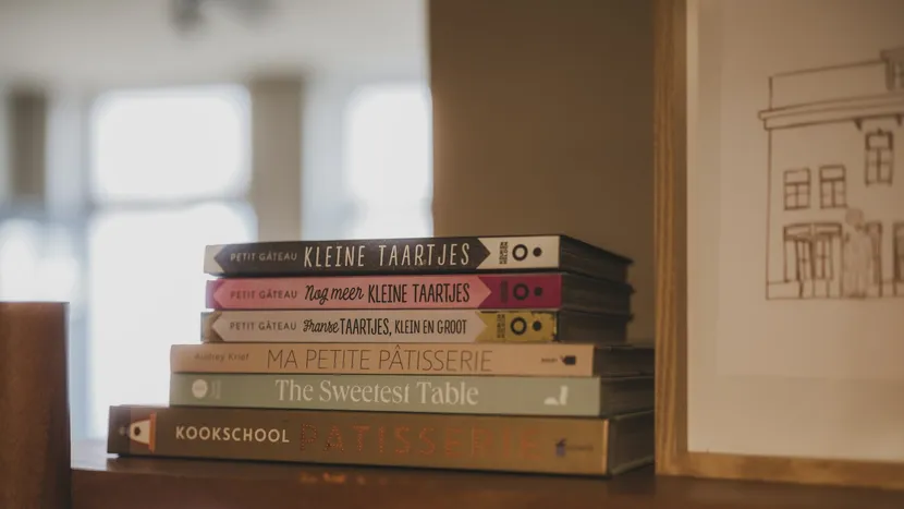 A stack of pâtisserie cookbooks on a wooden shelf by a window.