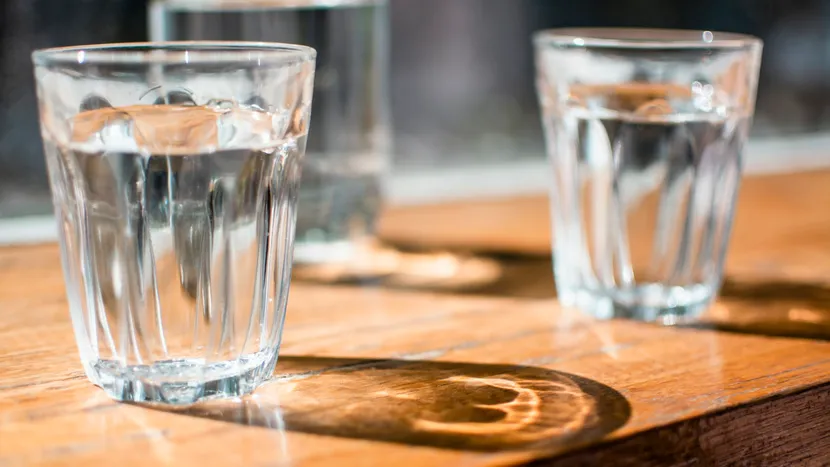 Two identical glasses of water on a sunlit wooden table.