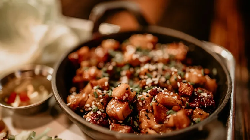Glazed cubes of pork with sesame seeds and chives in a small cast iron pan.