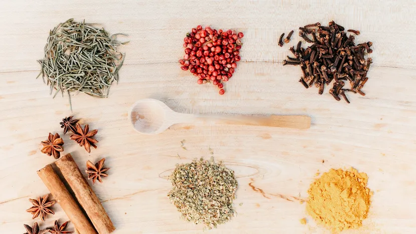Rosemary, pink peppercorns, cloves, star anise, cinnamon, oregano and turmeric laid out around a wooden spoon.