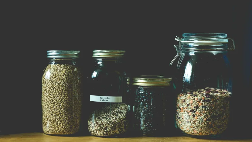 Four glass jars of grains, quinoa, black beans and mixed pulses on a wooden shelf, one of them labelled.