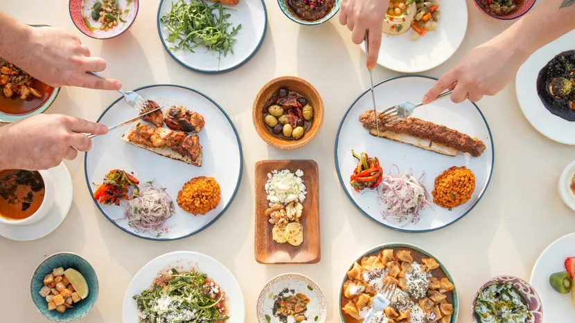 Seen from above, several hands reach across a table of shared plates, kebabs, olives and salads.