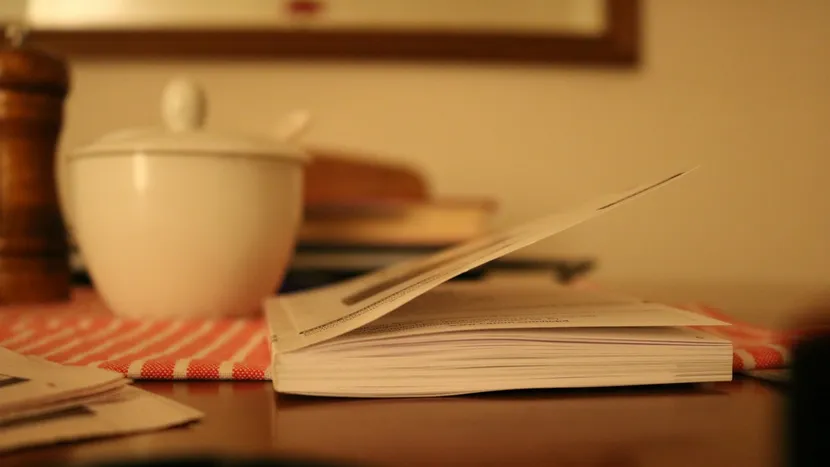 A paperback open on a kitchen table beside a sugar bowl and a striped dish towel.