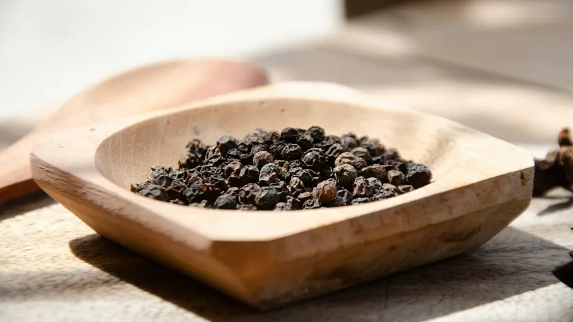 Whole black peppercorns in a carved wooden bowl, in warm sunlight.