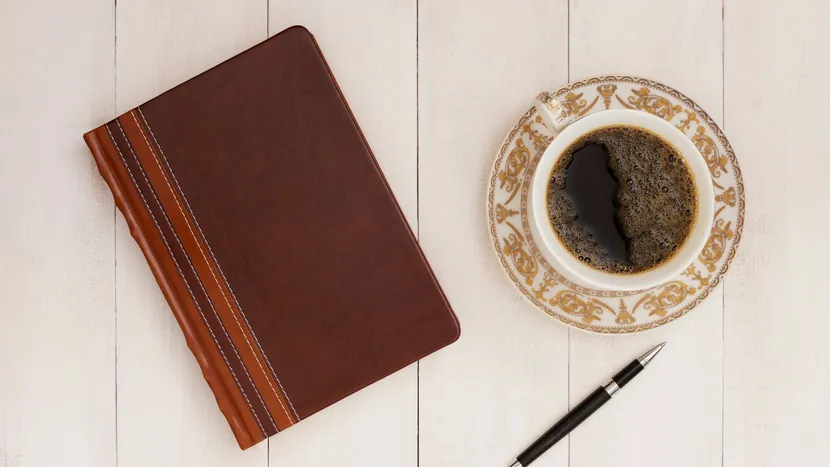 A closed leather notebook, a pen and a cup of coffee on white boards.