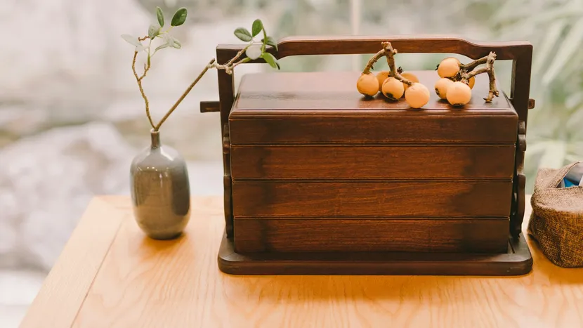 A tiered wooden food carrier with loquats on its lid, beside a small grey vase on a wooden table.